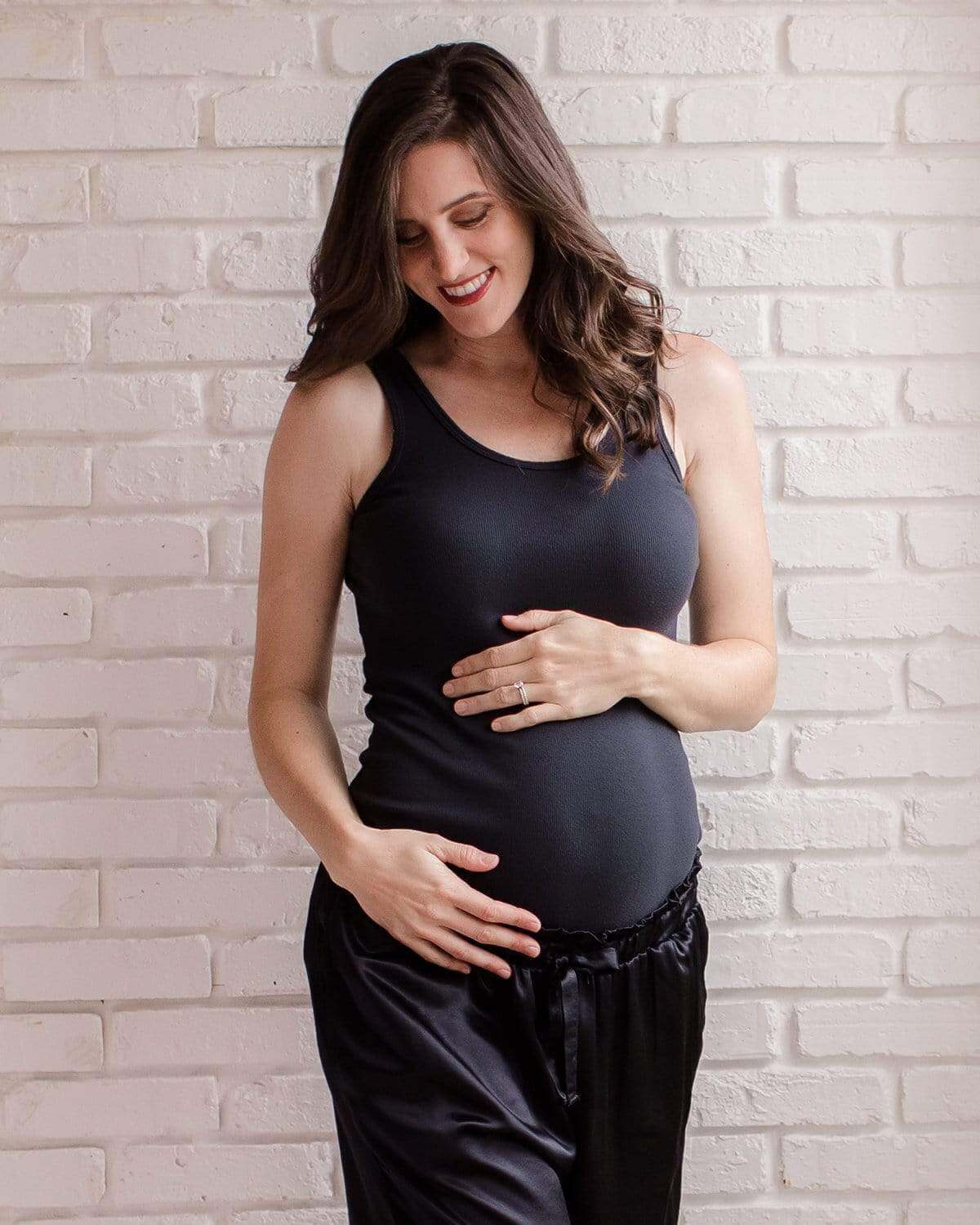 A pregnant woman wearing a beige tank top, sitting on a bed with a white brick wall in the background.