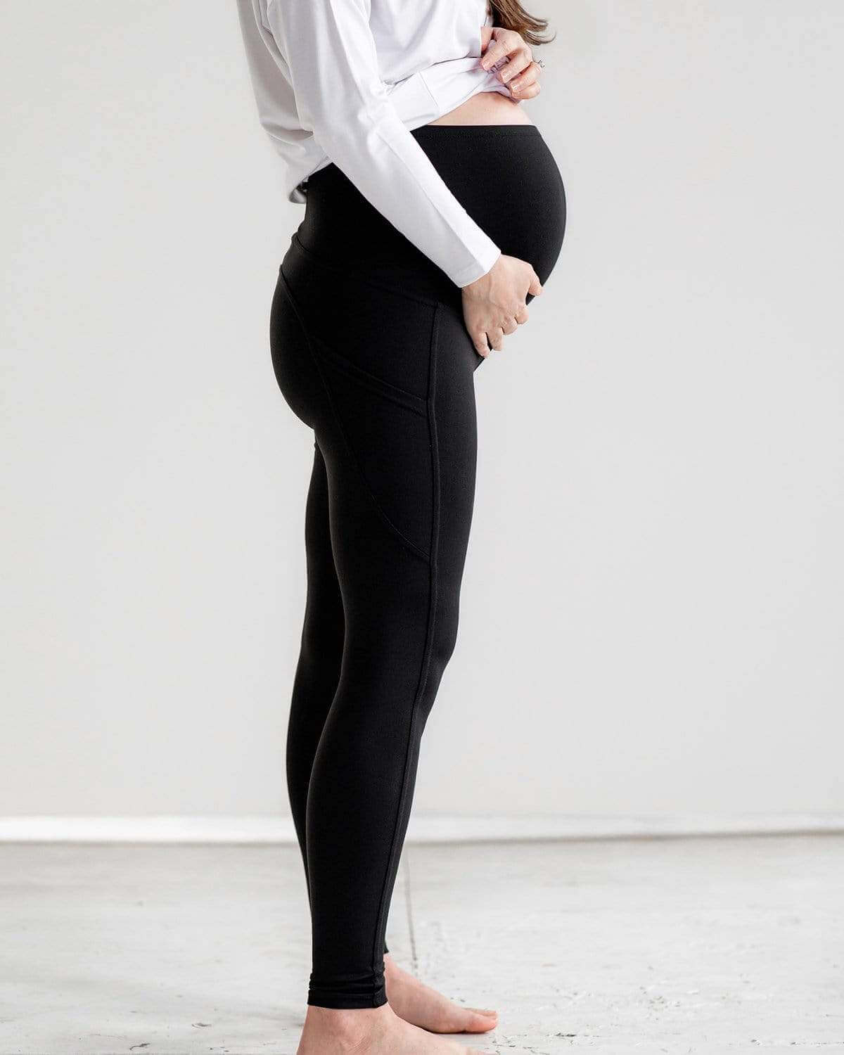 A pregnant woman wearing a white t-shirt and black maternity leggings with pockets, standing in a neutral setting.