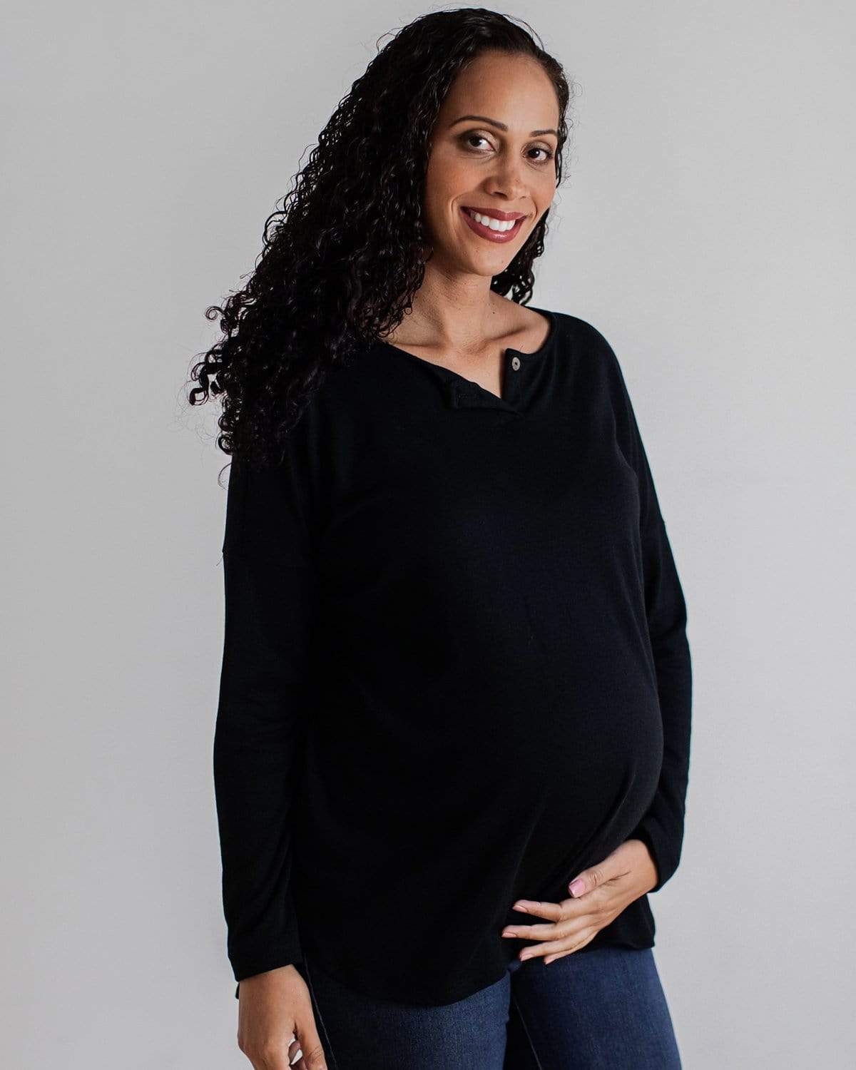 A pregnant woman wearing a black bamboo maternity henley with long dolman sleeves and a mini button placket.