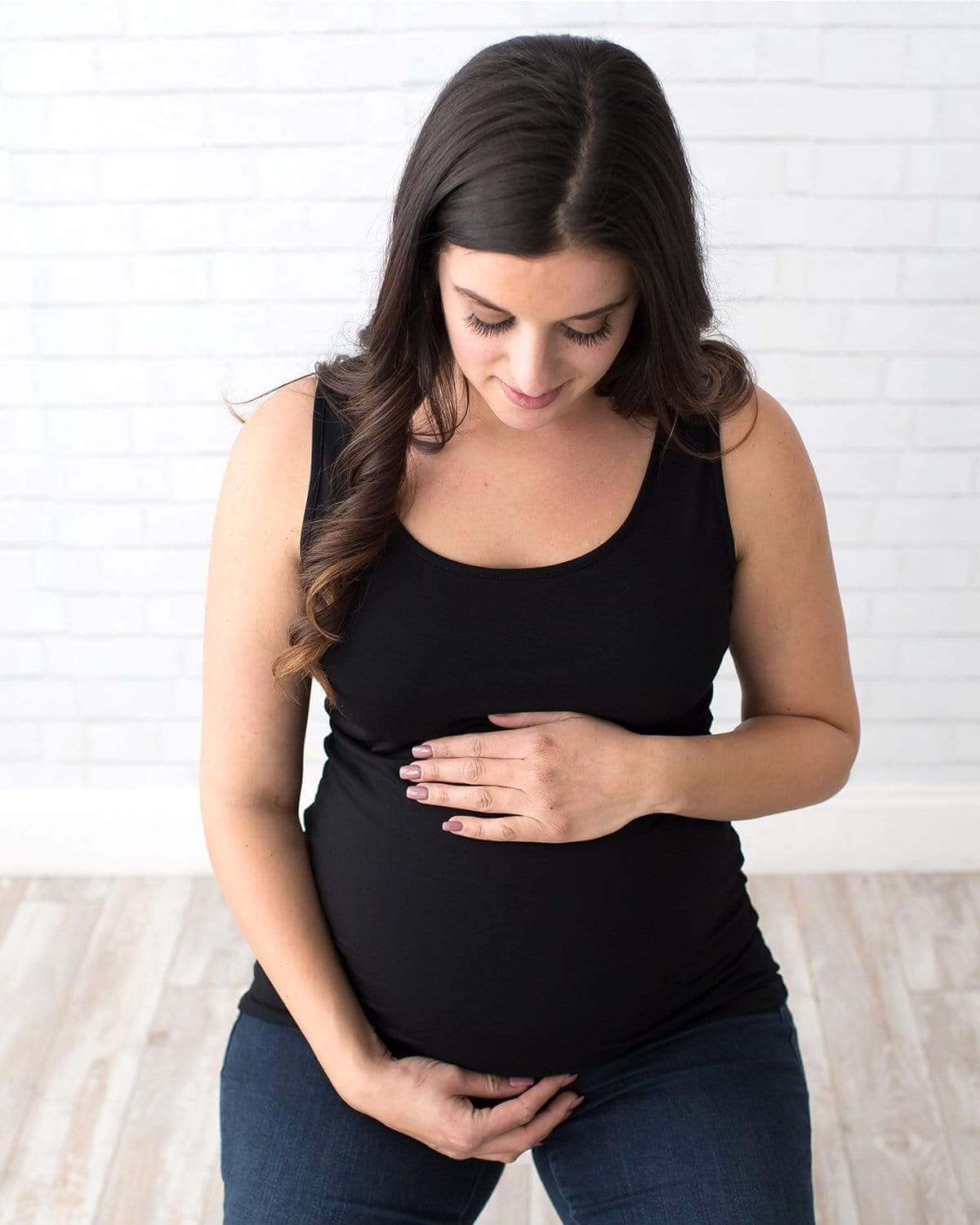 A pregnant woman wearing a white maternity tank top and blue jeans, standing in a relaxed pose.