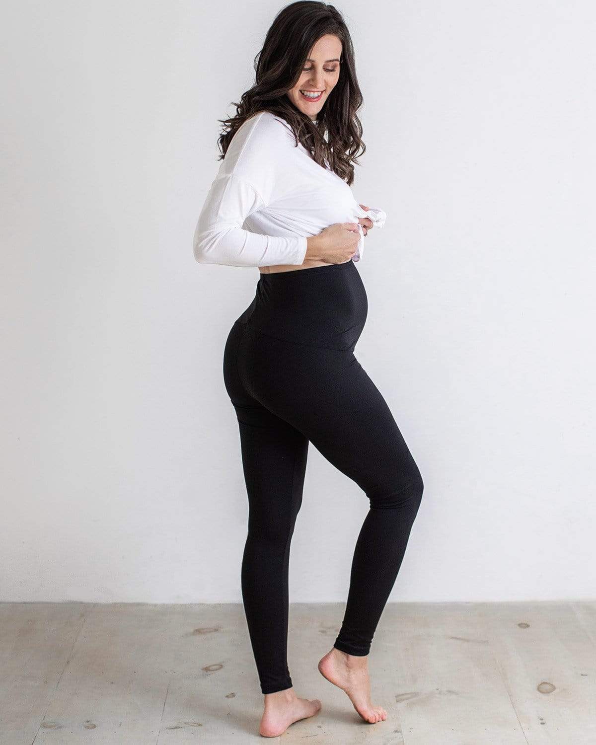A pregnant woman wearing black full-length maternity leggings with a comfort belly panel, paired with a white top.