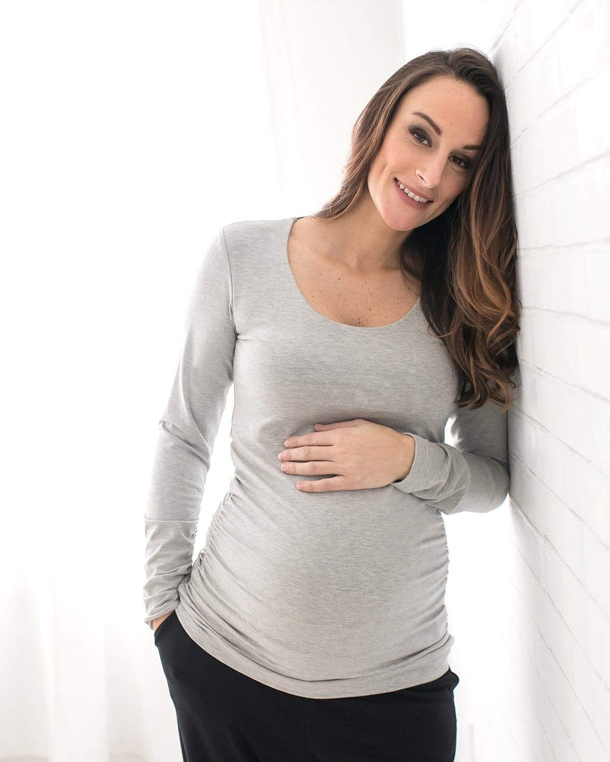A pregnant woman wearing a long sleeve black maternity top with a scoop neck, the top is ruched at the side, and she is sitting on a bench.
